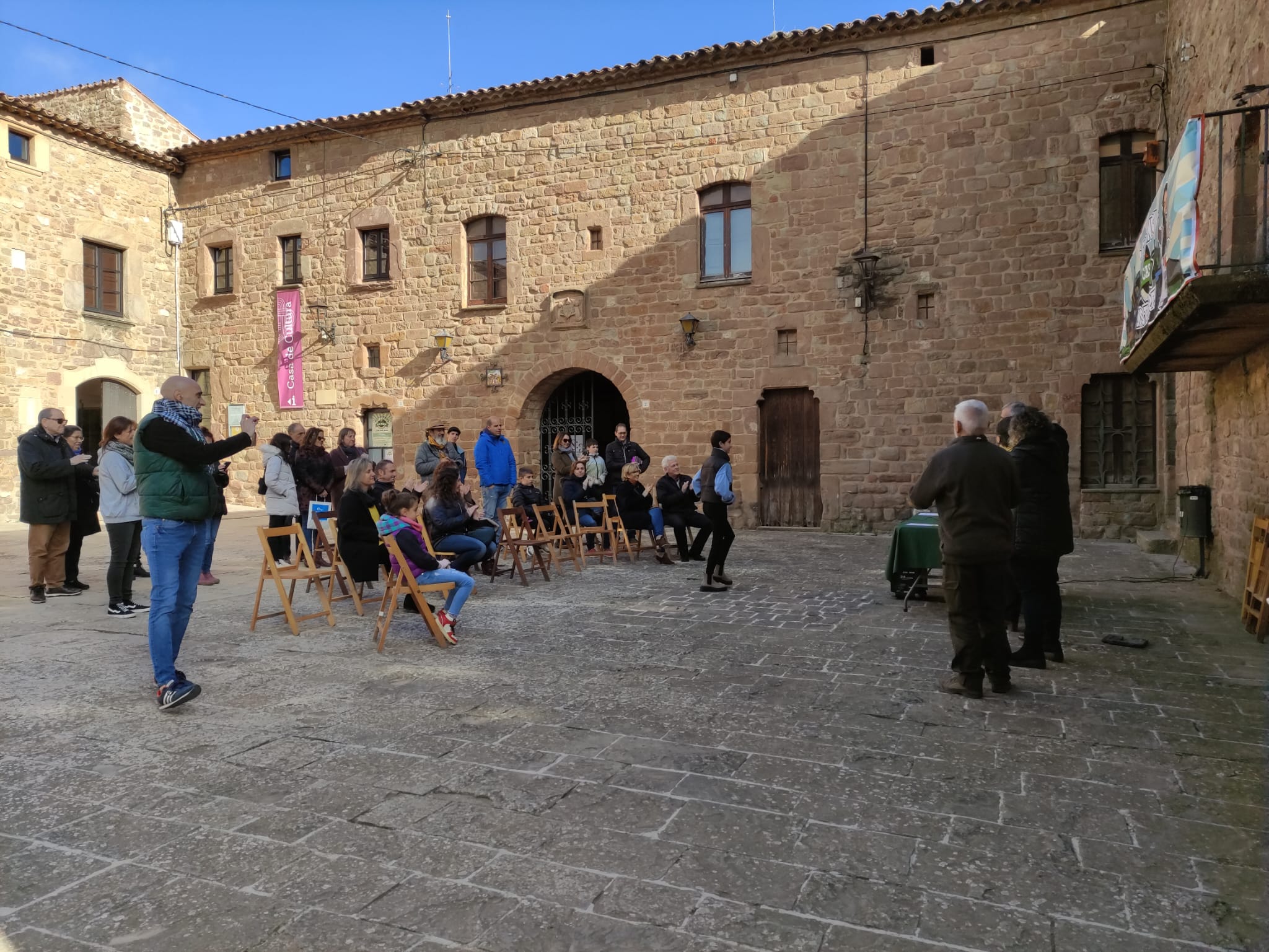 A la plaça del Monestir de l'Estan, un noi, que ha guanyat un dels premis dels jocs rurals va cap al taula on hi ha persones que representen als organitzadors i jurats per a recollir-lo. Es veu també part del públic assegut.