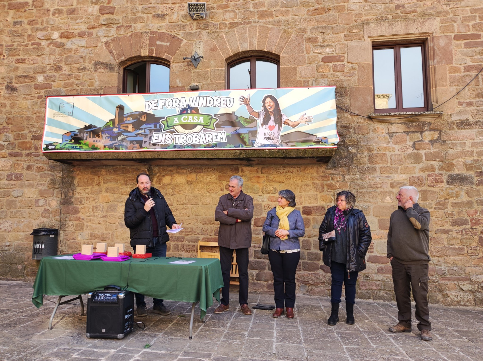 Imatge de cinc persones en un espai lliure darrera d'una taula en la que hi ha els guardons dels jocs florals que lliuraran a les persones premiades. Les cinc persones son Joan Solà, president de la sectorial de cultura de MdC, Mario Urrea president de MdC, Montse Barniol, delegada del governs a la Catalunya Central, Montse Isern, membre del jurat i Salvador Tresserra, alcalde de l'Estany la localiutat on es vam lliuar els premis dels Jocs Florals.